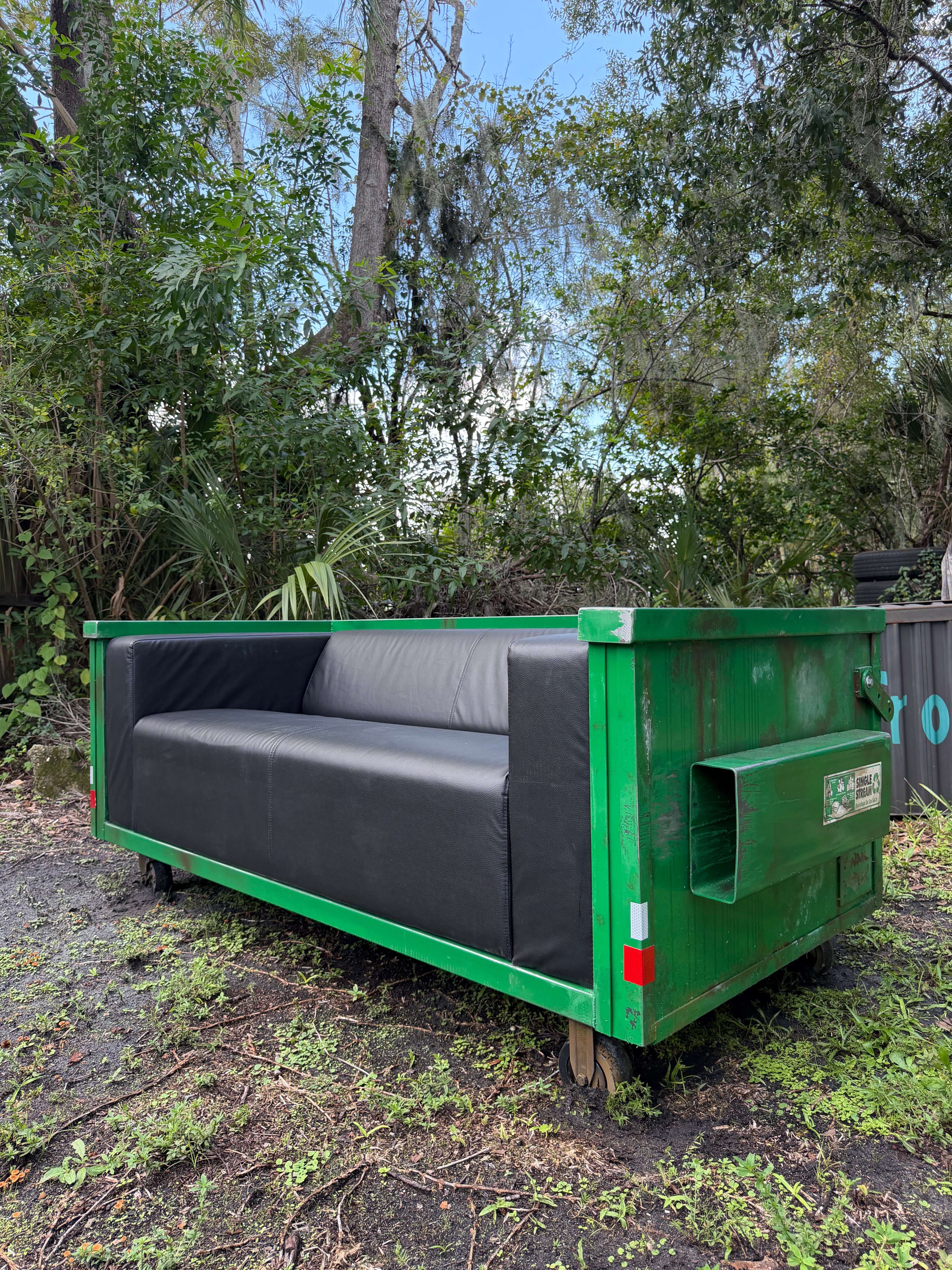 'Modular Waste' sofa by Ryder Studios x Gwak Productions, three-quarter front view: black vegan leather sofa set inside a hand-built green dumpster frame with fork pocket, reflectors and casters