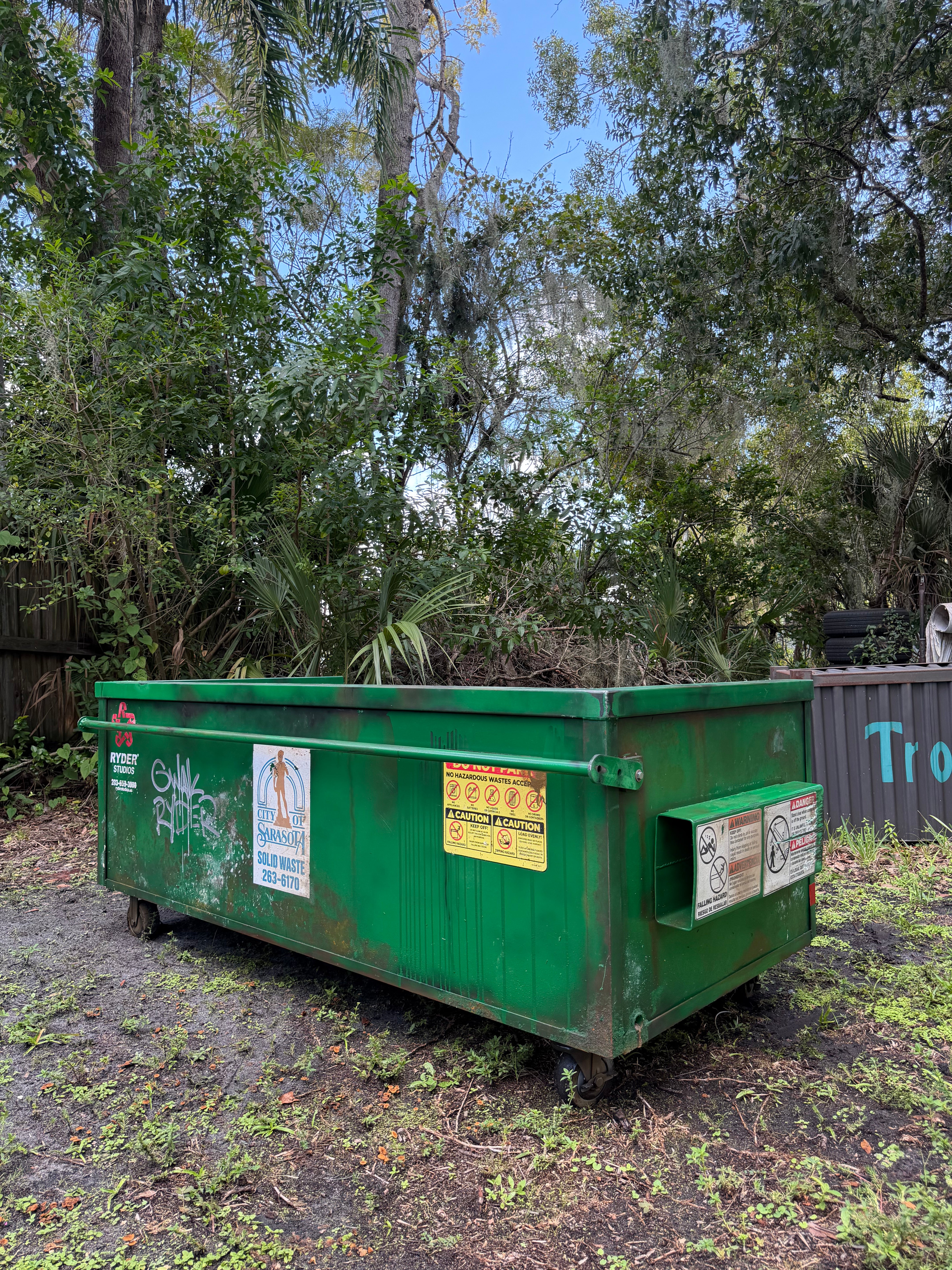 'Modular Waste' sofa, rear three-quarter view: green dumpster exterior with hazard stickers, side fork pocket and heavy-duty casters